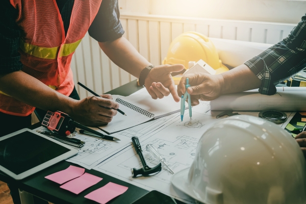Construction Project Planning Meeting with Safety Equipment Construction professionals reviewing blueprints and plans on desk with safety vest, hard hats, and tablet device during project planning meeting