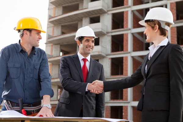 Construction manager in yellow hard hat, business executive in suit, and professional in white hard hat shaking hands at construction site with building framework in background
