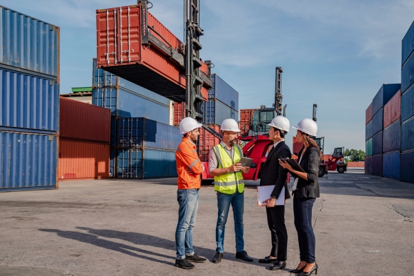 Shipping Container Yard Management and Logistics Planning Team of logistics professionals discussing operations in container terminal yard, wearing safety equipment and reviewing documents